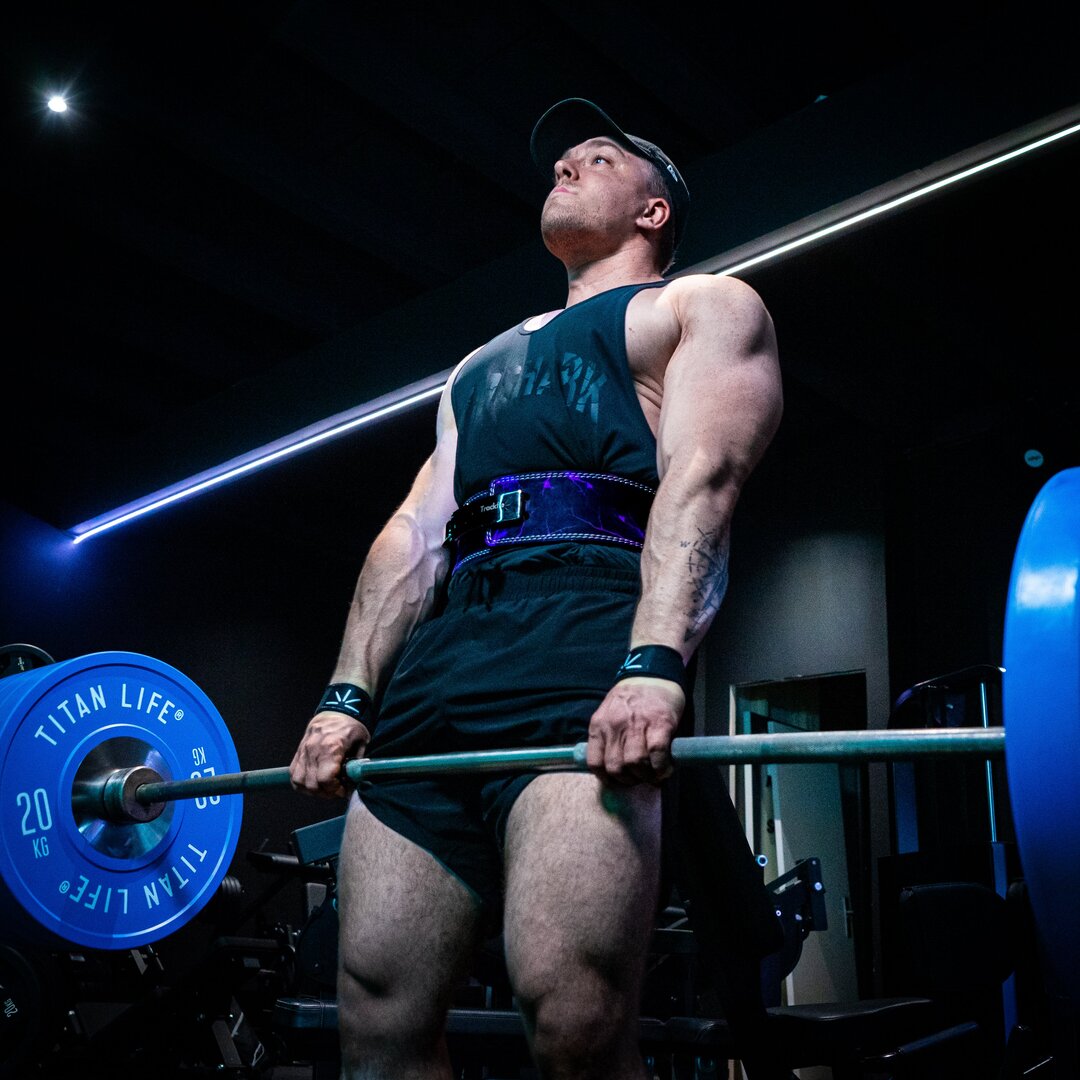 En mand dødløfter tungt og bruger lifting straps og et lilla træningsbælte i et fitnesscenter